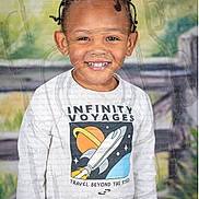 Xzavier is registered to the contest to win money with this photo: child, smiling, braided_hair, white_shirt, space_theme, rocket, outdoor, fence, greenery, portrait, happy, young, casual_clothing, standing, bright, colorful, cute, kid, fashion, nature