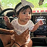 baby, child, infant, headband, bow, car, dashboard, drool, tattoo, hands, person, cute, curious, interior, window, greenery, seat, clothing, sitting, portrait