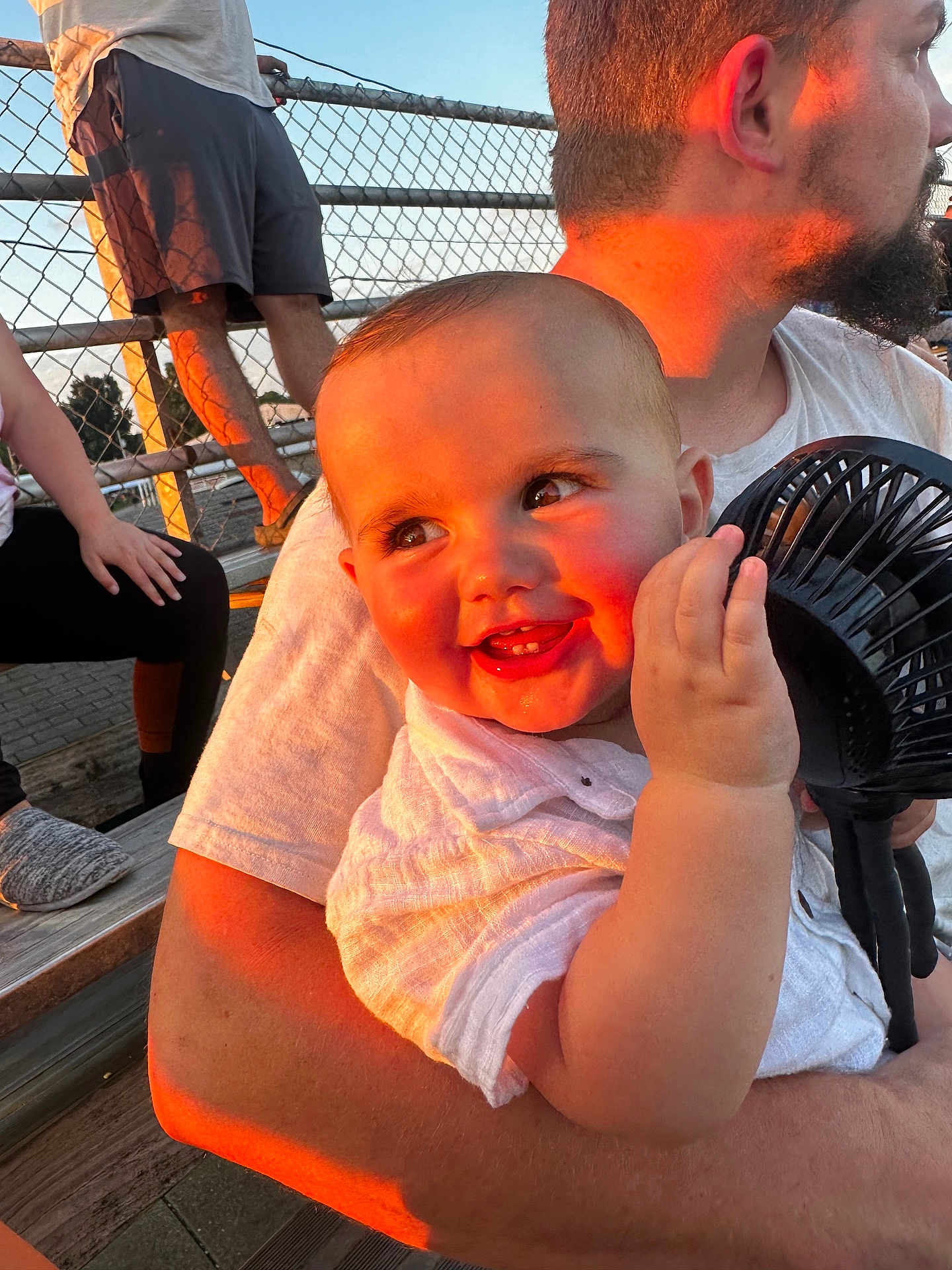 Brayden is registered to the contest to win money with this photo: baby, child, adult, smile, sunset, outdoor, fence, fan, white_clothing, person, beard, holding, bench, casual, happy, face, skin, hand, short_hair, light