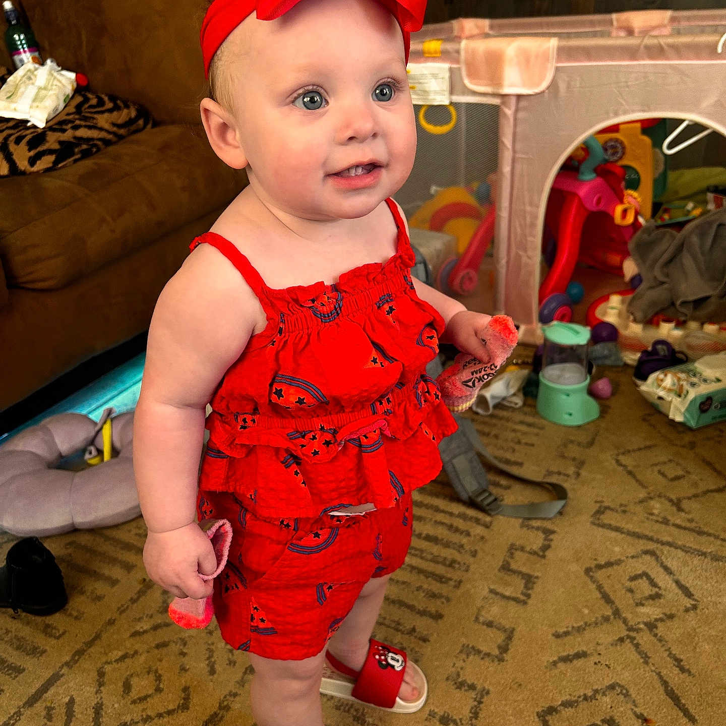 Everleigh is registered to the contest to win money with this photo: toddler, child, red_clothing, headband, indoor, carpet, toy, playpen, living_room, smiling, person, footwear, sock, baby, cute, standing, expression, home, furniture, play
