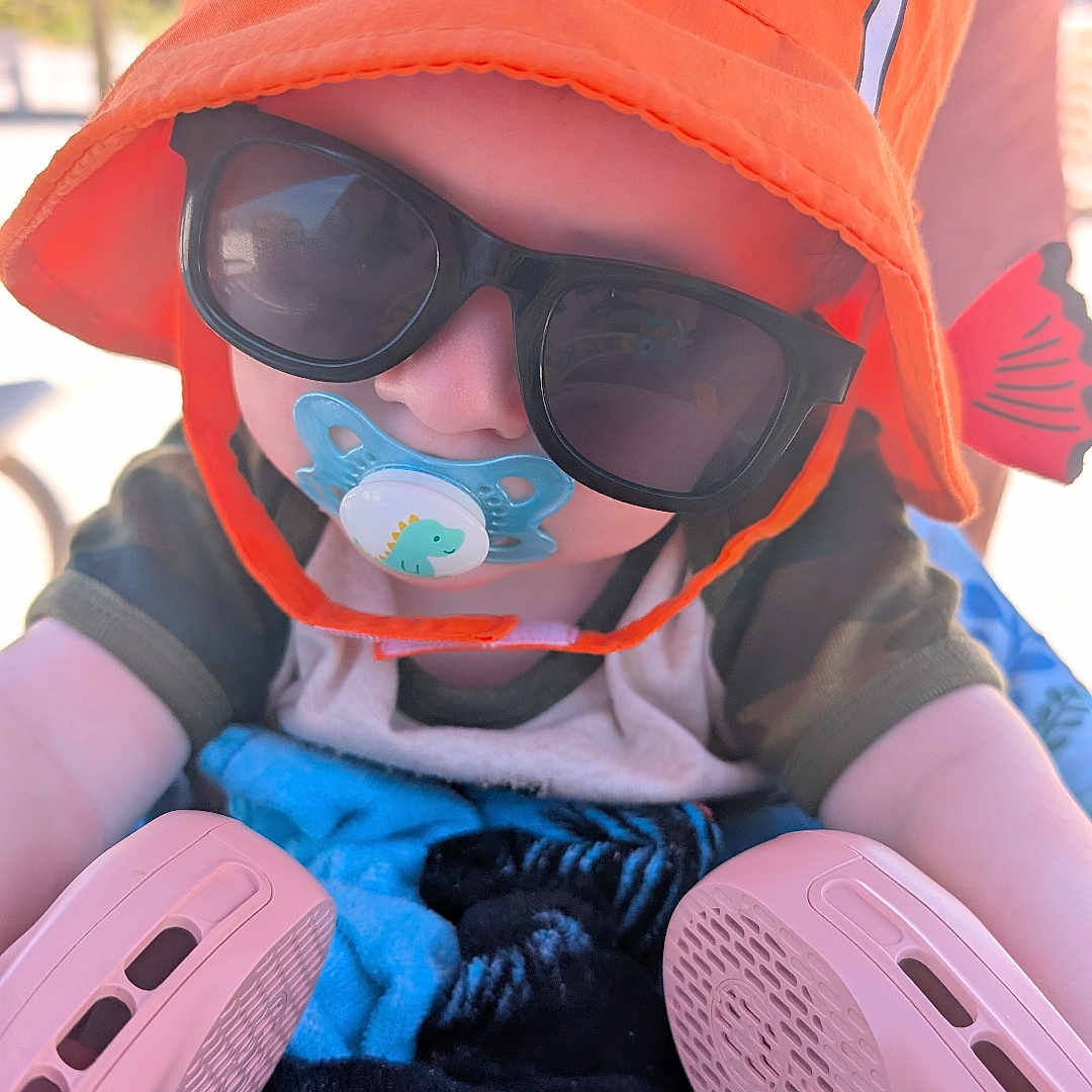 Murdock is registered to the contest to win money with this photo: accessory, baby, baby_walker, child, clothing, colorful, cute, face, hat, infant, orange_hat, outdoor, pacifier, person, playtime, summer, sunglasses, sunlight, toddler, toy