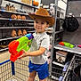 child, boy, cowboy_hat, toy_gun, shopping_cart, store, halloween_decorations, pumpkins, shelves, blue_shorts, gray_shirt, indoor, retail, playful, casual_clothing, smiling, person, footwear, plastic_toy, young