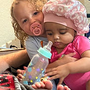 Meilani is registered to the contest to win money with this photo: baby, bottle, candid, child, closeup, curly_hair, cute, face, family, feet, hat, holding, indoor, infant, pacifier, person, pink_clothing, sitting, toddler, young_child