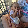 baby, child, person, adult, hands, bottle, thumb_sucking, curly_hair, earrings, clothing, porch, sunlight, shadow, chair, lap, nail_polish, relaxing, casual, home, outdoor