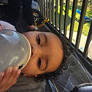Meilani is registered to the contest to win money with this photo: toddler, baby, bottle, milk, curly_hair, face, hand, sunlight, balcony, railing, outdoor, person, child, relaxing, drinking, casual, daylight, clothing, background, floor
