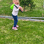 Gennevieve is registered to the contest to win money with this photo: child, girl, young_child, playing, toy_golf_club, sneakers, pink_shoes, jeans, long_sleeve_shirt, chainlink_fence, grass, lawn, outdoor, tree, backyard, portrait, casual_clothing, daylight, hair_tied, curious