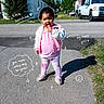 Nova is registered to the contest to win money with this photo: toddler, child, pink_clothing, lollipop, outdoor, sunny_day, doodle_art, text_overlay, shadow, driveway, grass, building, vehicle, smiling, happy, person, casual_wear, curly_hair, footwear, playful
