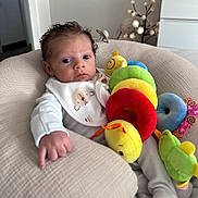 Riven is registered to the contest to win money with this photo: baby, infant, plush_toy, caterpillar_toy, bib, cushion, indoor, soft_lighting, stuffed_animal, furniture, child, person, cute, face, hand, white_clothing, relaxing, portrait, home, decor