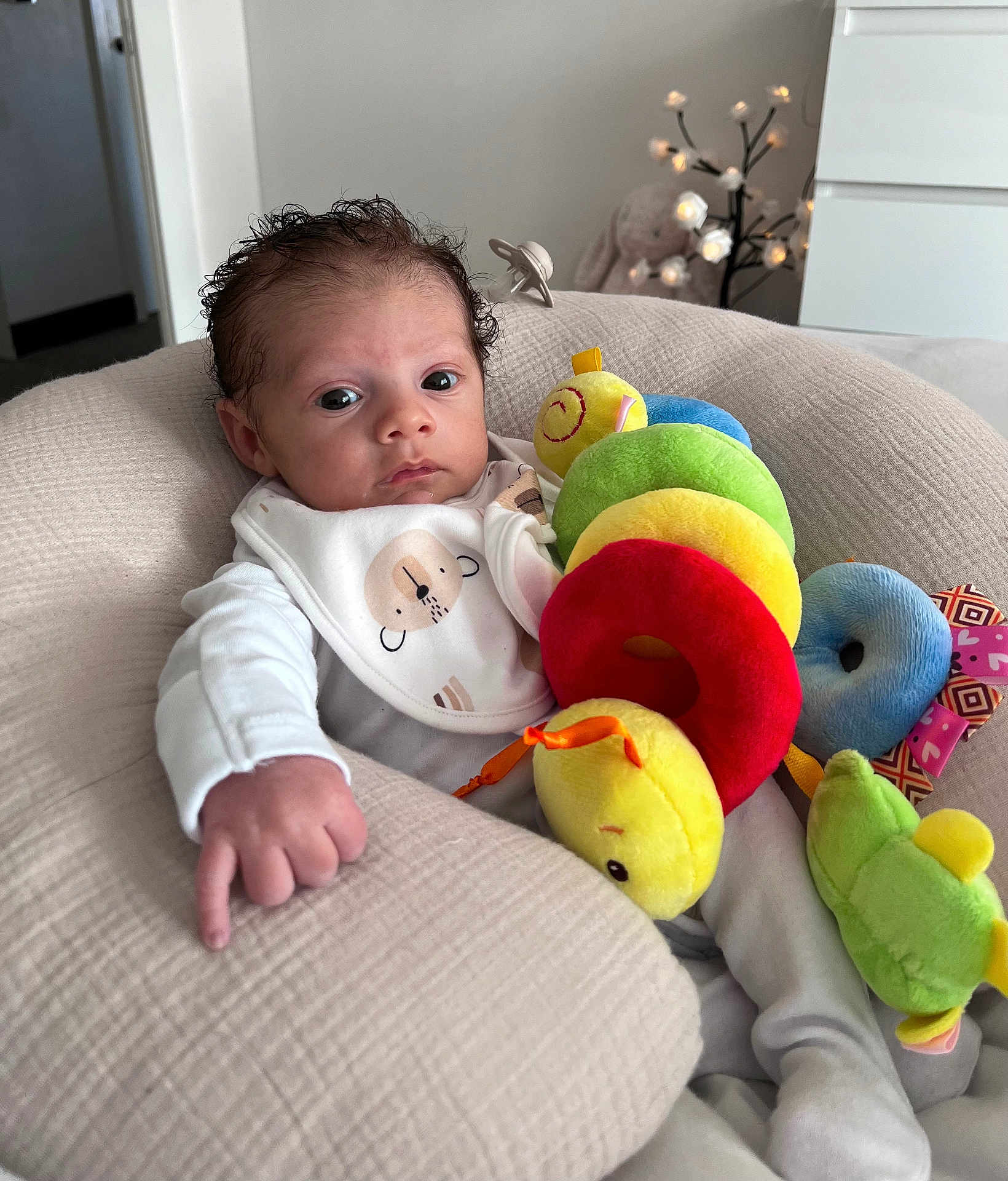 Riven is registered to the contest to win money with this photo: baby, infant, plush_toy, caterpillar_toy, bib, cushion, indoor, soft_lighting, stuffed_animal, furniture, child, person, cute, face, hand, white_clothing, relaxing, portrait, home, decor