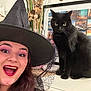 person, cat, black_cat, witch_hat, smiling, makeup, purple_eyeshadow, red_lipstick, long_hair, indoor, cabinet, decor, dried_plants, picture_frame, cityscape, feline, pet, happy, costume, portrait