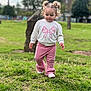 Charlotte is registered to the contest to win money with this photo: building, casual_clothing, child, curly_hair, cute, daylight, grass, greenery, nature, outdoor, park, pigtails, pink_pants, portrait, stone, toddler, trees, walking, white_sweatshirt, young_child
