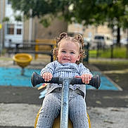 Charlotte is registered to the contest to win money with this photo: toddler, child, playground, spring_rider, curly_hair, pigtails, grey_clothing, knitwear, socks, shoes, outdoor, daylight, play, happy, smile, park, bench, fence, trees, background_blur