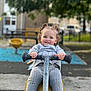toddler, child, playground, spring_rider, curly_hair, pigtails, grey_clothing, knitwear, socks, shoes, outdoor, daylight, play, happy, smile, park, bench, fence, trees, background_blur