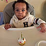 toddler, cupcake, birthday_candle, high_chair, celebration, child, curly_hair, white_jacket, table, party_decorations, colorful_streamers, indoor, face, hands, expression, food, festive, young_child, cute, person