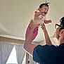 baby, child, adult, indoor, curtains, window, smile, glasses, person, joy, hair, lifting, playful, pink_clothing, white_shirt, room, furniture, light, hands, happy