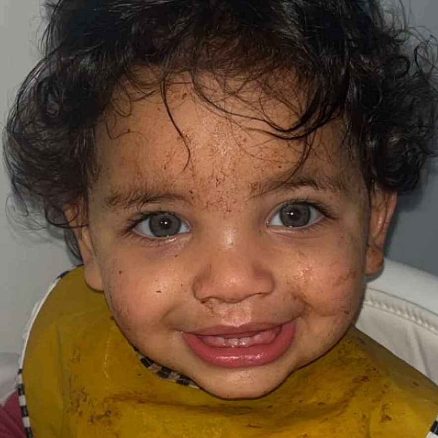 Sofia participe au concours pour gagner de l'argent avec cette photo : toddler, child, curly_hair, blue_eyes, smiling, messy_face, food_stains, yellow_bib, highchair, indoor, portrait, happy, cute, clothing, person, young_child, baby, mealtime, fun, expression