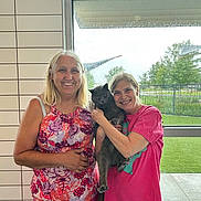 My Son is registered to the contest to win money with this photo: two_women, cat, indoor, window, rain, smiling, gray_cat, casual_clothing, floral_pattern, pink_shirt, plaid_pants, holding_pet, human_face, daylight, fence, green_grass, portrait, happy, female, natural_light
