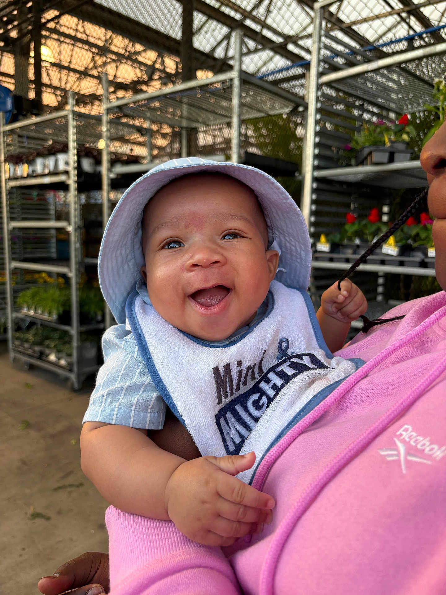Brian Welch joined the competition — help win amazing prizes! baby, smiling, child, bib, hat, bucket_hat, blue_hat, person, holding, pink_hoodie, clothing, plant_nursery, plants, metal_racks, indoor, happy, portrait, closeup, face, joyful