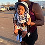 Brian Welch is registered to the contest to win money with this photo: baby, adult, sunhat, bib, pacifier, outdoor, parking_lot, person, child, holding, shorts, sleeveless_top, blue_socks, afternoon_light, shadow, vehicle, curious, casual_clothing, closeup, portrait
