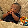 baby, child, couch, snack, cheetos, puffed_snack, beige, clothing, furniture, indoor, person, portrait, sitting, young_child, curly_hair, dark_skin, hand, face, relaxation, thinking