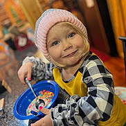 Kinsley is registered to the contest to win money with this photo: child, toddler, smiling, hat, pink_hat, cereal, bowl, spoon, milk, breakfast, kitchen, countertop, granite, yellow_shirt, checked_shirt, indoor, person, food, happy, cute