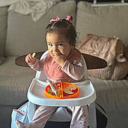 Danielle is registered to the contest to win money with this photo: toddler, child, high_chair, food, snack, pink_bow, hair_accessory, bib, pajamas, indoor, living_room, sofa, plate, spoon, cute, baby, person, feeding, seated, smiling