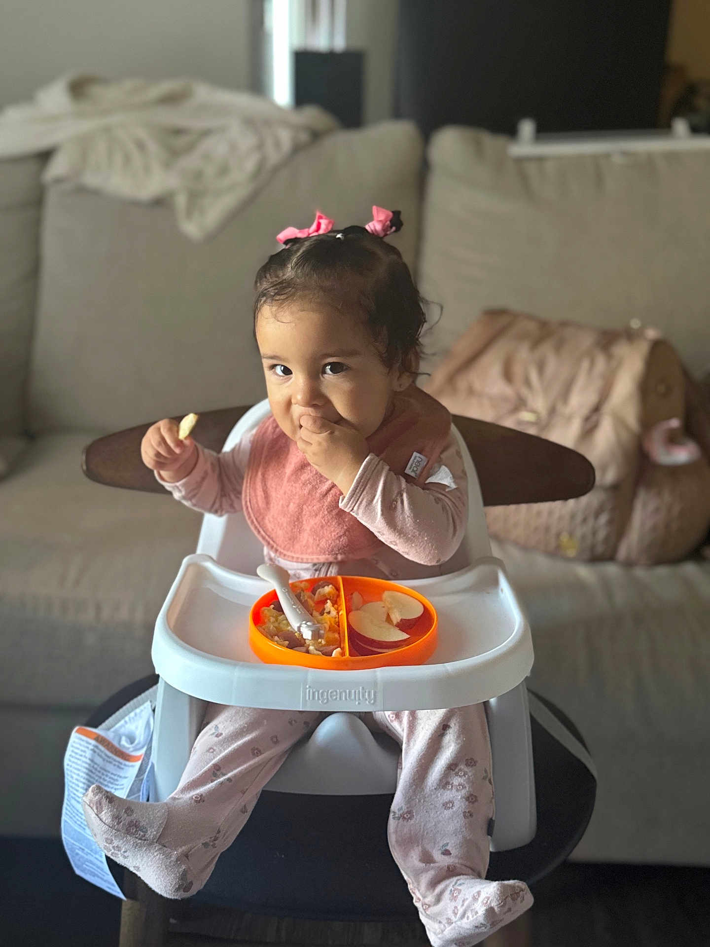 Danielle is registered to the contest to win money with this photo: toddler, child, high_chair, food, snack, pink_bow, hair_accessory, bib, pajamas, indoor, living_room, sofa, plate, spoon, cute, baby, person, feeding, seated, smiling