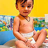 baby, bare_chest, brown_eyes, changing_pad, child, closeup, colorful_background, crossed_legs, dark_hair, diaper, happy, indoors, necklace, nursery, playroom, portrait, sitting, smiling, toddler, wall_art