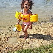 Serena a rejoint le concours — aidez-le/la à gagner de superbes lots ! child, curly_hair, floaties, yellow, bucket, lake, water, sand, shore, grass, sunny, smiling, barefoot, outdoor, play, summer, nature, recreation, happy, person