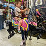 Noah joined the competition — help win amazing prizes! child, adult, carousel, carousel_horse, amusement_ride, carnival, fair, lights, canopy, stuffed_toy, smile, holding, metal_floor, shoe, sock, shorts, pole, colorful, crowd, portrait