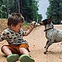 child, toddler, boy, dog, pet, stuffed_toy, dirt_road, outdoors, trees, forest, play, candid, smiling, sitting, shorts, patterned_shirt, dinosaur_print, green_shoes, collar, interaction