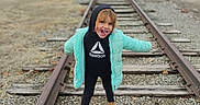 Lani is registered to the contest to win money with this photo: child, railroad_tracks, jacket, boots, outdoor, playful, tongue_out, gravel, wooden_sleepers, overcast, person, young_child, casual_clothing, nature, smiling, fun, winter_clothing, standing, hoodie, expression