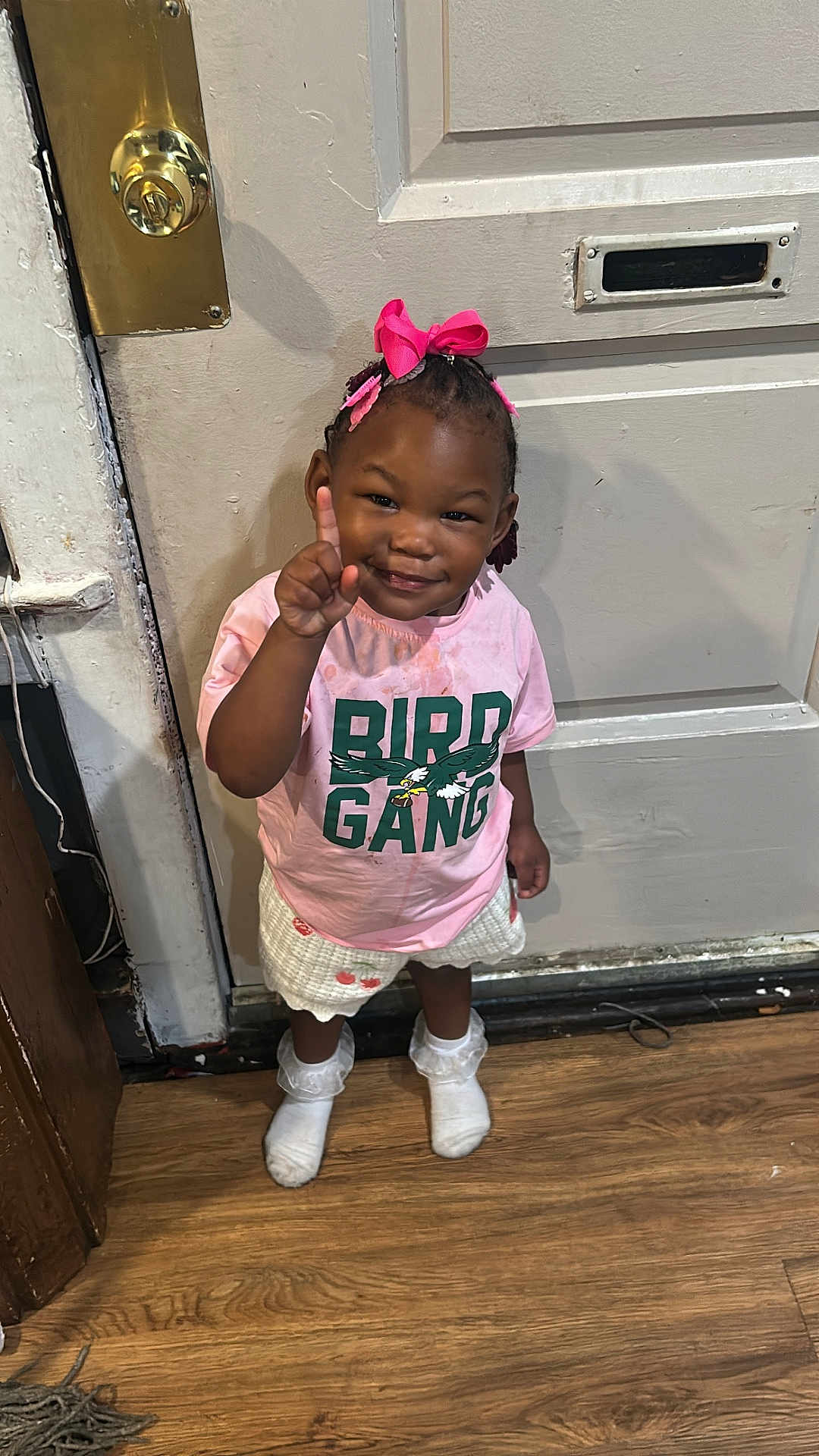 Milan is registered to the contest to win money with this photo: toddler, child, girl, indoor, wooden_floor, pink_bow, hair_accessory, tshirt, shorts, socks, smiling, pointing, door, doorknob, floor, clothing, person, cute, standing, happy