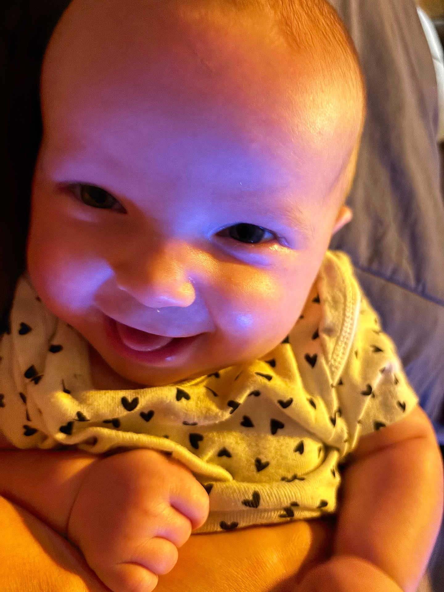 Bailee Rockholt is registered to the contest to win money with this photo: baby, infant, smiling, close_up, onesie, hearts_pattern, eyes, cheeks, hand, fist, skin, head, portrait, adorable, happy, candid, colorful_lighting, purple_tint, blanket, lying_down