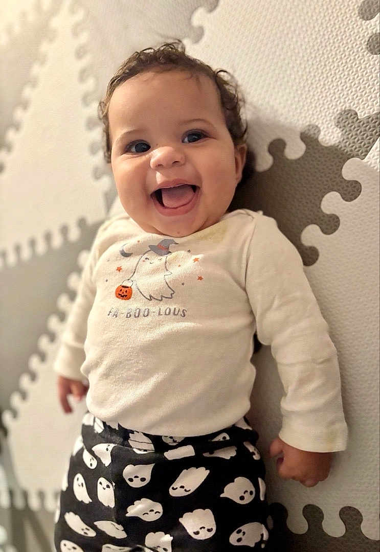 Cataleya is registered to the contest to win money with this photo: baby, child, smiling, happy, face, curly_hair, long_sleeve_shirt, halloween, ghost_print, puzzle_mat, play_mat, infant, cute, indoors, portrait, clothing, toddler, person, expression, fun