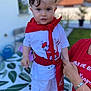 toddler, child, red_scarf, white_shirt, white_shorts, outdoor, person, hand, adult_support, blurred_background, greenery, patterned_tablecloth, short_hair, cute, standing, clothing, casual, portrait, daylight, expression