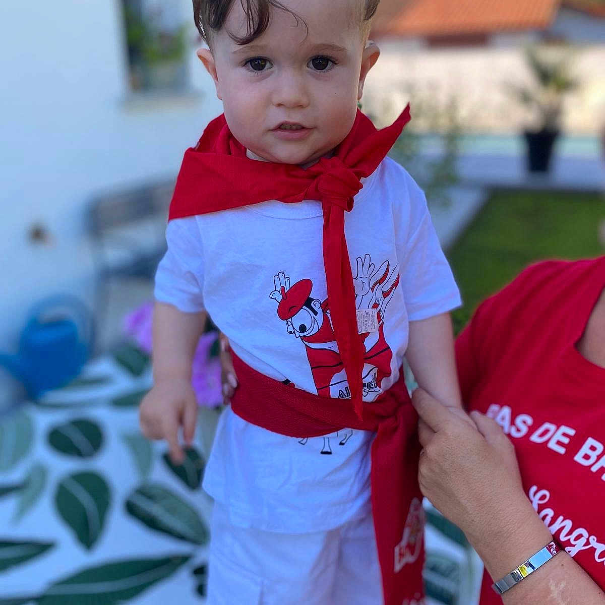Bixente participe au concours pour gagner de l'argent avec cette photo : adult_support, blurred_background, casual, child, clothing, cute, daylight, expression, greenery, hand, outdoor, patterned_tablecloth, person, portrait, red_scarf, short_hair, standing, toddler, white_shirt, white_shorts