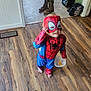 child, costume, spiderman, mask, snack, barefoot, floor, wood_floor, plastic_container, indoor, boots, backpack, door, light, smile, food, candid, home, playful, young_child