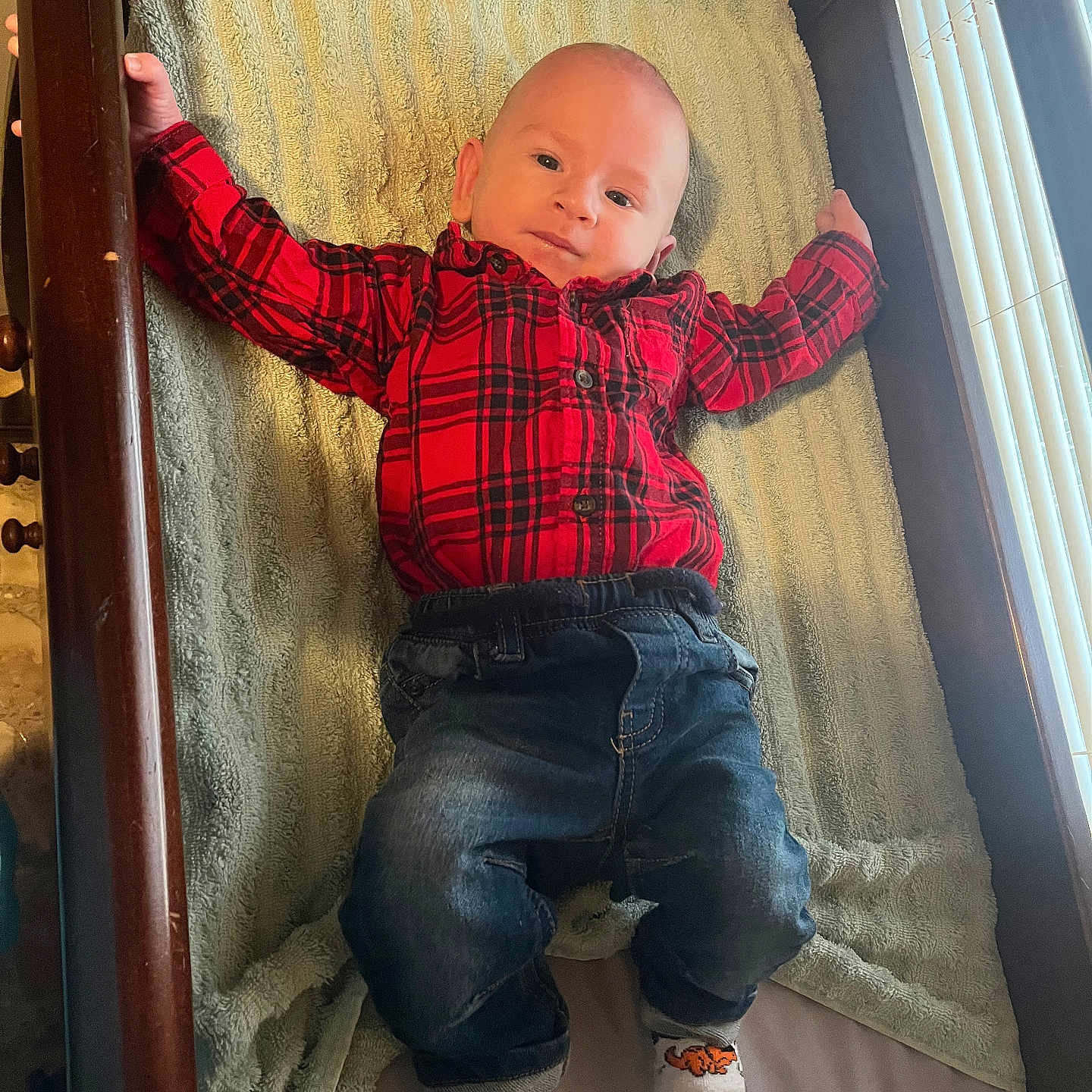 Wyatt is registered to the contest to win money with this photo: baby, child, plaid_shirt, jeans, socks, crib, blanket, wooden_crib, indoor, portrait, smiling, infant, cute, relaxed, person, baby_clothing, soft_texture, comfortable, casual, resting