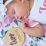 newborn, baby, sleeping, infant, portrait, close_up, headband, flower, blanket, floral, pink, white, onesie, arm, hand, face, peaceful, milestone, wooden_sign, indoor