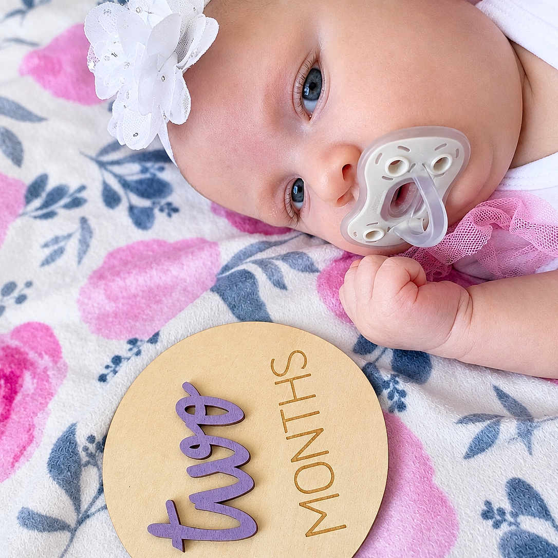 Lila is registered to the contest to win money with this photo: baby, infant, portrait, lying_down, pacifier, flower_headband, white_clothing, pink_clothing, fist, floral_blanket, wooden_sign, milestone_photo, two_months, face, eyes, indoors, close_up, nursery, cute, baby_photography