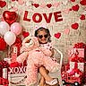child, toddler, teddy_bear, heart_shaped_sunglasses, pink_bow, white_chair, valentine_decor, red_hearts, pink_hearts, balloons, gift_boxes, wooden_floor, smiling, indoor, cute, love_banner, pink_shoes, toy_truck, celebration, happy