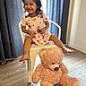 child, toddler, smiling, sitting, chair, red_bow, dress, hearts, sunglasses, teddy_bear, plush_toy, indoors, curtains, wooden_floor, carpet, happy, cute, playful, person, portrait