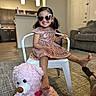 Melani Garcia is registered to the contest to win money with this photo: child, girl, heart_shaped_sunglasses, pink_dress, sparkly_clothing, white_chair, teddy_bear, pink_teddy_bear, living_room, carpet, sofa, couch, wooden_floor, indoor, smiling, pigtails, happy, playful, furniture, foot