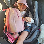 Maëlyne participe au concours pour gagner de l'argent avec cette photo : child, car_seat, backpack, pink_hat, smiling, seatbelt, car_interior, window, sunlight, casual_clothing, happy, young_girl, blue_shorts, holding, sitting, seat, safety, travel, person, kid