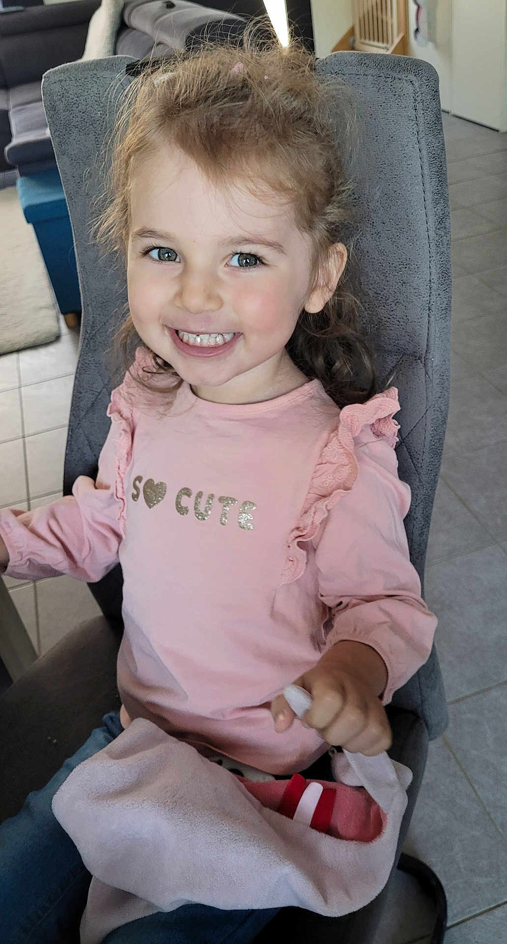 Maëlyne a rejoint le concours — aidez-le/la à gagner de superbes lots ! child, girl, smiling, pink_shirt, blanket, chair, indoor, happy, portrait, cute, casual_clothing, toddler, home, furniture, person, long_hair, holding, seated, tile_floor, cozy