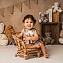toddler, child, smiling, wicker_chair, teddy_bear, plush_toy, brown, cream, curly_hair, cute, happy, sitting, wooden_crate, bunting_flags, indoor, portrait, baby_clothes, floor, decor, cozy