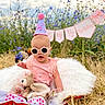 baby, sunglasses, party_hat, birthday, outdoor, flowers, teddy_bear, blanket, grass, celebration, child, cute, plush_toy, nature, field, pink_clothing, sunny, smiling, portrait, fun