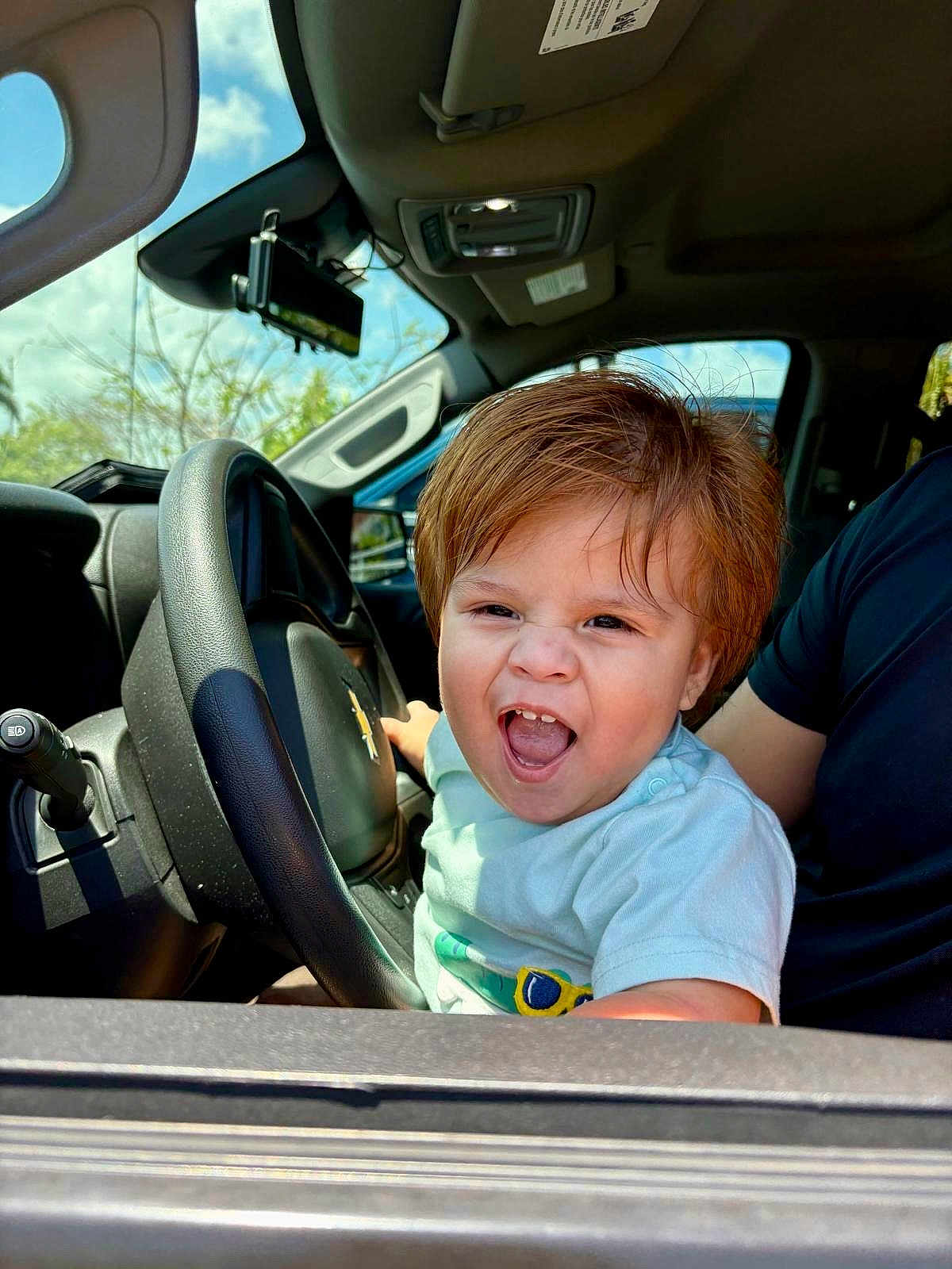 Harvey Ruiz joined the competition — help win amazing prizes! toddler, child, car, steering_wheel, interior, daylight, happy, excited, person, short_sleeves, seat, automobile, young_child, hair, smile, open_mouth, window, dashboard, seatbelt, adult_arm