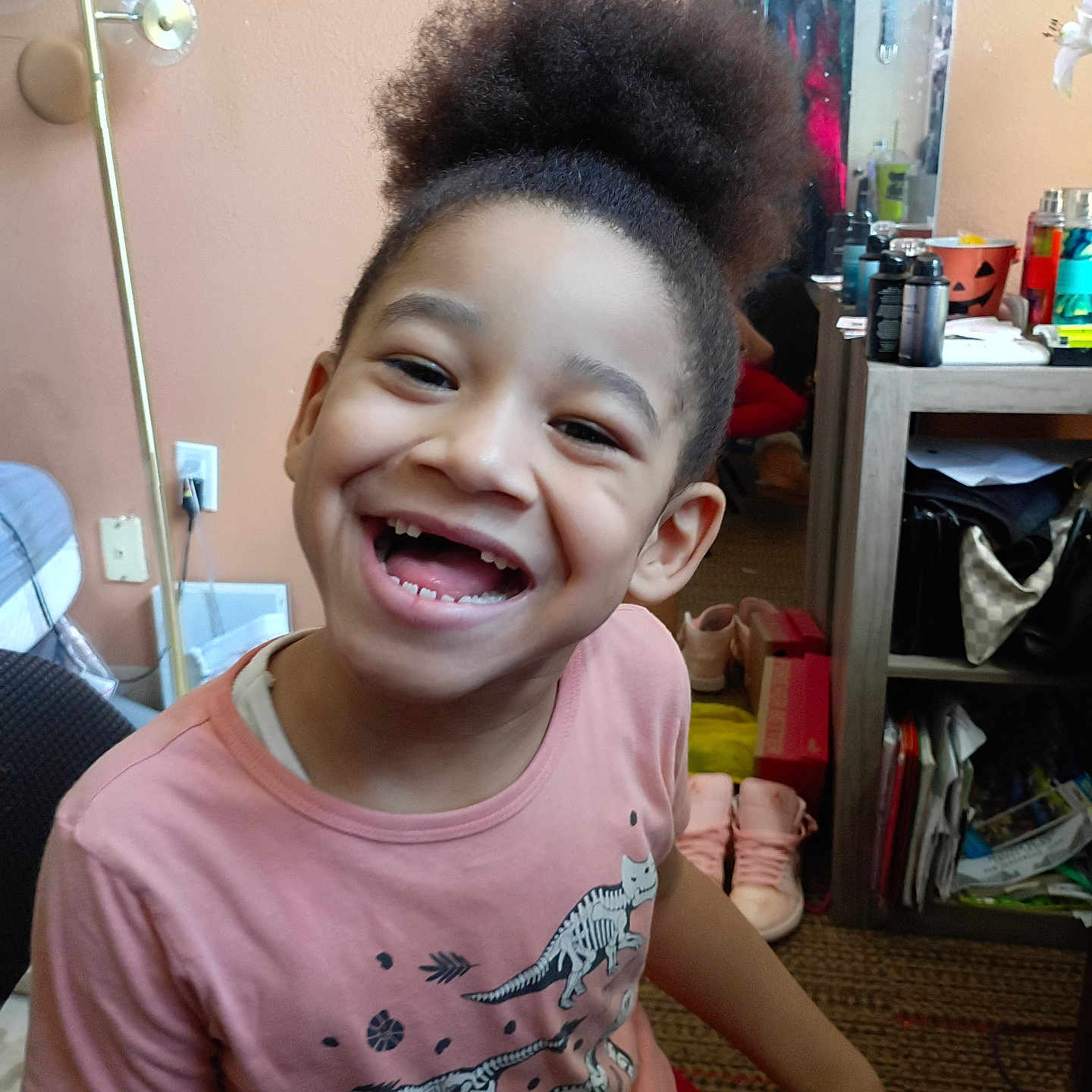 Jonas is registered to the contest to win money with this photo: child, smiling, happy, indoor, controller, game_controller, person, casual_clothing, pink_shirt, hair_puff, room, mirror, shoes, sitting, furniture, clutter, home, toy, portrait, joyful